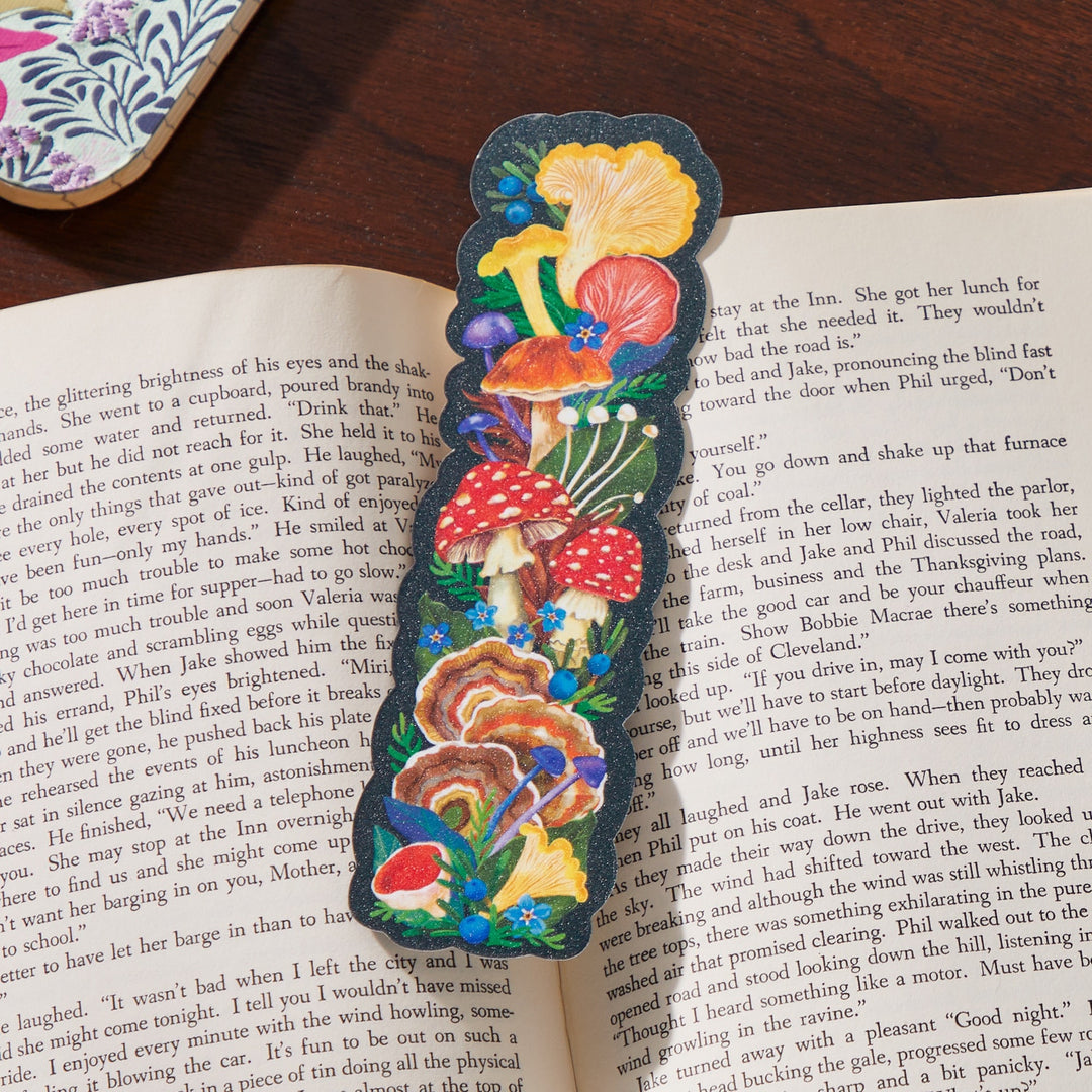 The World of Mushrooms Paper Bookmark by Olga Akbarova showcases vibrant red, yellow, and brown mushrooms with green leaves and blue flowers on a striking black background, adding a colorful touch to any open book.