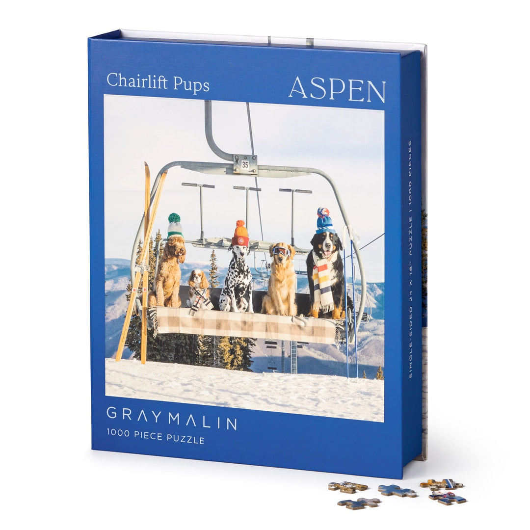 The Gray Malin Aspen 1000 Piece Book Box Puzzle features fine art photography of four dogs in winter hats on a ski lift over snowy mountains. The puzzle comes in a box with the title and brand, with some pieces shown nearby.