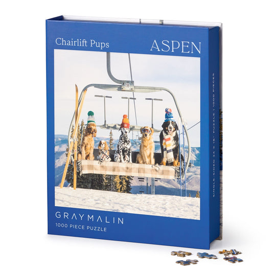 The Gray Malin Aspen 1000 Piece Book Box Puzzle features fine art photography of four dogs in winter hats on a ski lift over snowy mountains. The puzzle comes in a box with the title and brand, with some pieces shown nearby.