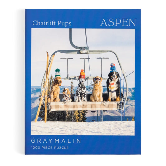 The Gray Malin Aspen 1000 Piece Book Box Puzzle shows five winter-clad dogs on a ski lift with snowy mountains, featuring Gray Malin’s Aspen photography. The box reads “Gray Malin 1000 Piece Puzzle.”.