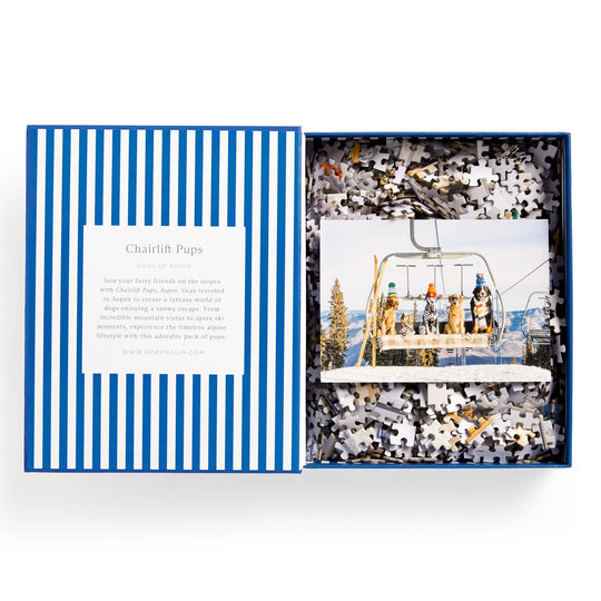 The Gray Malin Aspen 1000 Piece Book Box Puzzle by Gray Malin features a photo of four dogs on a ski lift amid snowy trees, titled “Chairlift Pups.” The open box displays blue and white stripes and loose puzzle pieces.
