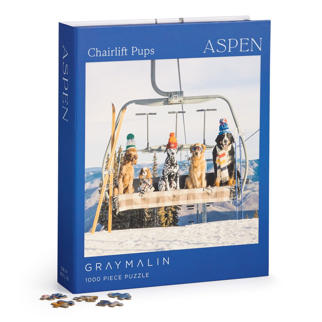 The Gray Malin Aspen 1000 Piece Book Box Puzzle by Gray Malin features five dogs in winter hats and scarves sitting on a ski lift above snowy mountains, showcasing his fine art photography.