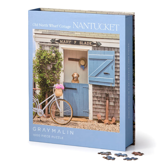 The Gray Malin Nantucket 1000 Piece Book Box Puzzle by Gray Malin features a coastal cottage with weathered shingles, blue Dutch door, dog peeking out, and pink flowers on a bicycle—ideal for Nantucket photography fans.