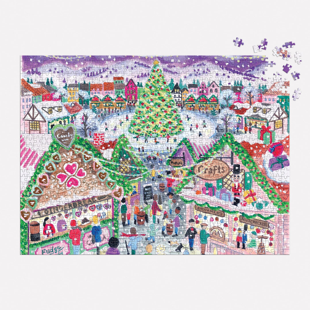 The Michael Storrings Holiday Village Market 1000pc Foil Puzzle features a festive winter village scene with a large Christmas tree, snow-covered rooftops, market stalls, and people shopping amid falling snow.