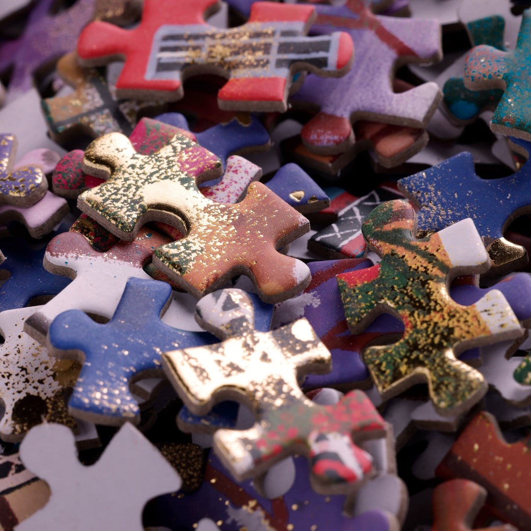 A close-up of Joy Laforme Twinkletown Express 1000 Piece Foil Puzzle pieces, showcasing vibrant colors, gold glitter accents, and unique painted patterns from the Joy Laforme brand.