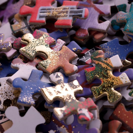 A close-up of Joy Laforme Twinkletown Express 1000 Piece Foil Puzzle pieces, showcasing vibrant colors, gold glitter accents, and unique painted patterns from the Joy Laforme brand.