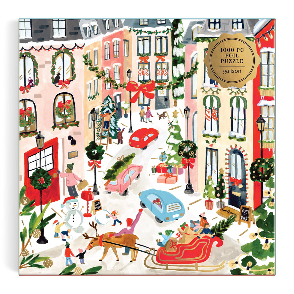 Festive city street puzzle features decorated buildings, people, a dog-pulled sleigh, gifts, Christmas trees, and cars; gold label in the corner reads “Merriest Village 1000 Piece Foil Puzzle Sabina Fenn.”.