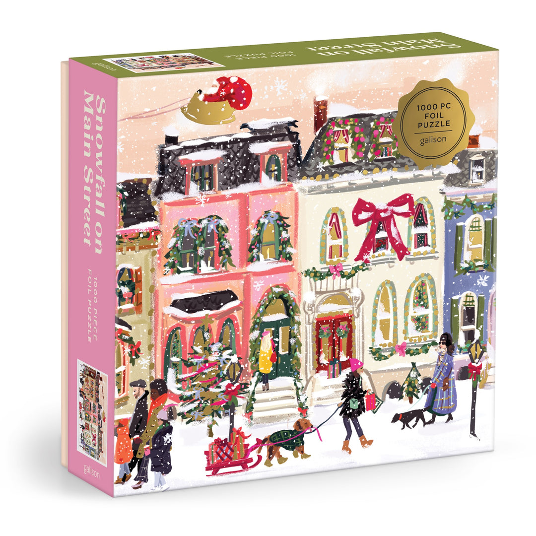The Snowfall on Main Street 1000 Piece Foil Puzzle by Nysha Lilly showcases a festive, snowy main street with colorful houses, children playing, and shimmering foil accents highlighting holiday décor and the sparkling Christmas tree.