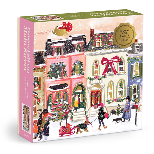 The Snowfall on Main Street 1000 Piece Foil Puzzle by Nysha Lilly showcases a festive, snowy main street with colorful houses, children playing, and shimmering foil accents highlighting holiday décor and the sparkling Christmas tree.