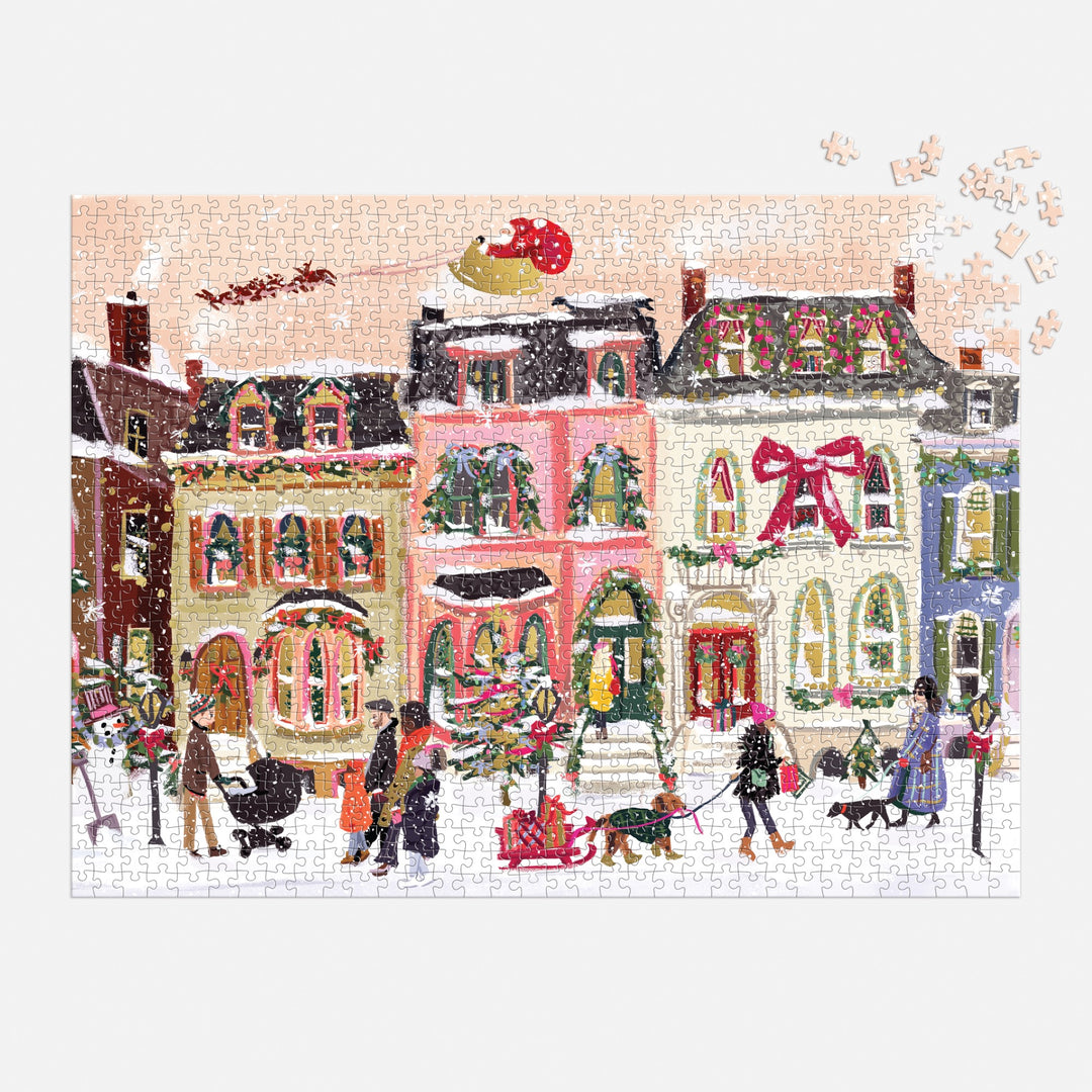 Nysha Lilly’s Snowfall on Main Street 1000 Piece Foil Puzzle showcases a festive winter street with decorated houses, children playing, and Santa’s sleigh overhead—just a few sparkling foil pieces remain to complete it.