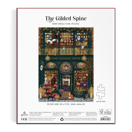 The Gilded Spine 1000 Piece Foil Puzzle by Sally Wilson features a cozy bookstore scene with bookshelves, hanging lights, readers, and a cat by the door. Finished puzzle measures 20 x 27 inches.