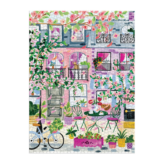 The Cherry Lane Blossoms 1000 Piece Puzzle by Sabina Fenn features a vibrant building with pink and purple walls, balconies adorned with plants, flowering trees, a bicycle, and an outdoor table with chairs among potted flowers.