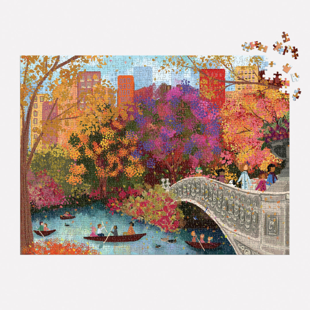 The Joy Laforme Autumn on Bow Bridge 1000 Piece Puzzle features a vibrant fall scene of Central Park’s Bow Bridge, people boating, and city buildings. Most of the puzzle is assembled with a few pieces missing from the top right corner.
