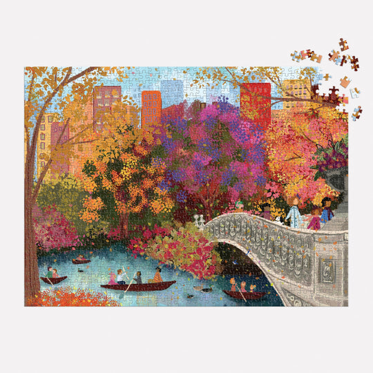 The Joy Laforme Autumn on Bow Bridge 1000 Piece Puzzle features a vibrant fall scene of Central Park’s Bow Bridge, people boating, and city buildings. Most of the puzzle is assembled with a few pieces missing from the top right corner.