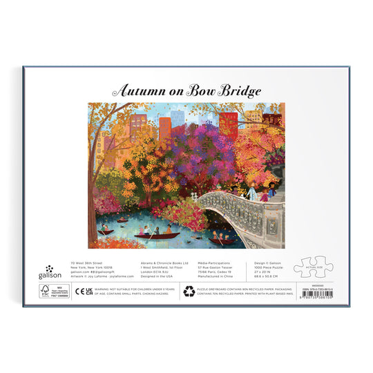 The Joy Laforme Autumn on Bow Bridge 1000 Piece Puzzle displays an autumn scene of Central Park’s Bow Bridge, colorful trees, and boaters, with “Autumn on Bow Bridge” above the illustration and Joy Laforme branding below.