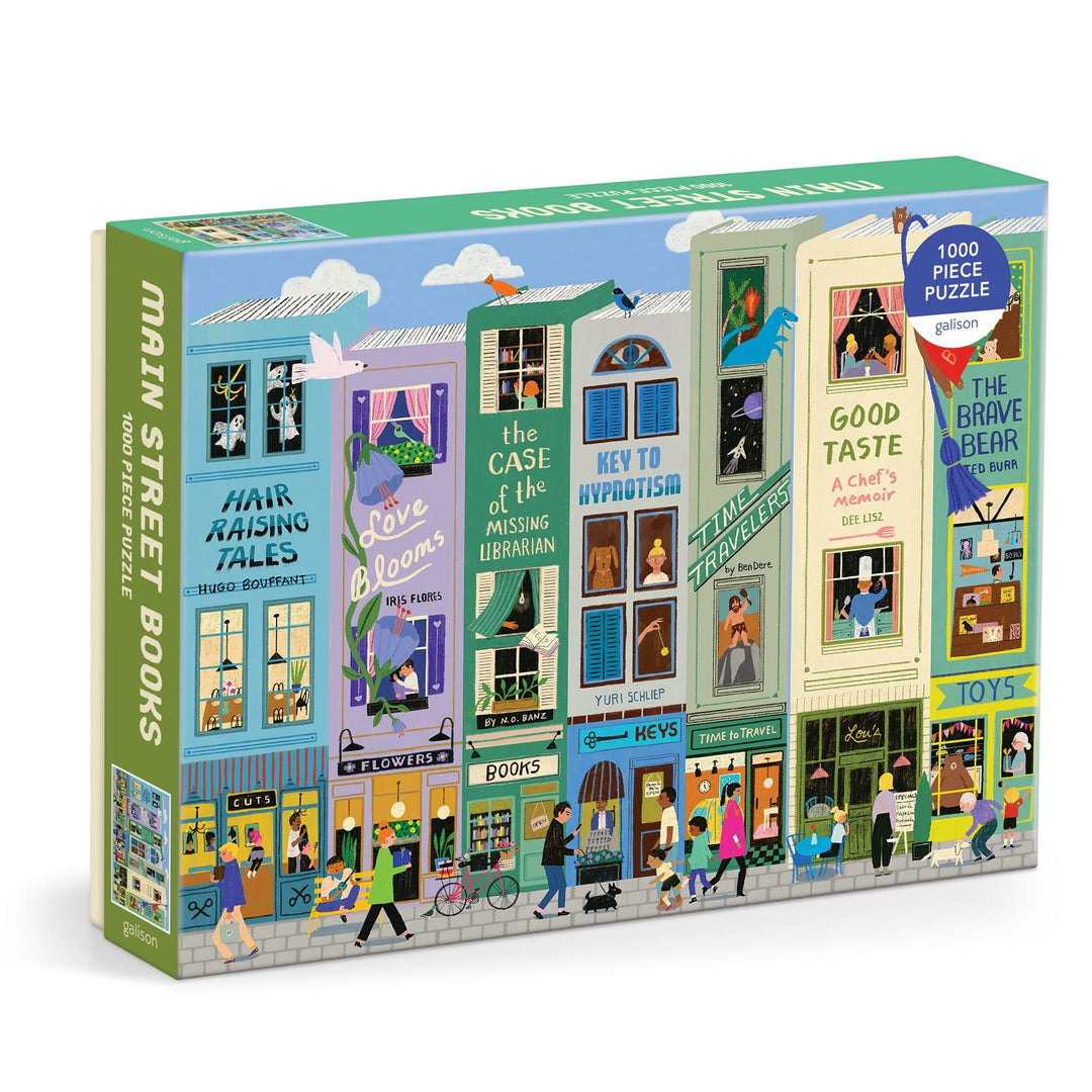 The Anne Bentley Main Street Books 1000 Piece Puzzle features illustrated whimsical bookshop storefronts, each with unique signs, books, flowers, people, a blue sky with birds, and a cat on a rooftop.
