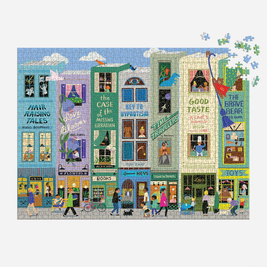 The Main Street Books 1000 Piece Puzzle by Anne Bentley features a vibrant street of playful bookshops, whimsical signs, and pedestrians. A green dragon pops out of a shop window, though a few pieces are missing in the top right corner.
