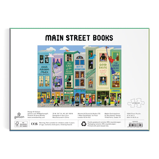 Illustration of a vibrant street with shops and apartments, each storefront showing playful book-themed signs. People stroll, bike, and read on the bustling sidewalk. Text: Main Street Books 1000 Piece Puzzle by Anne Bentley.