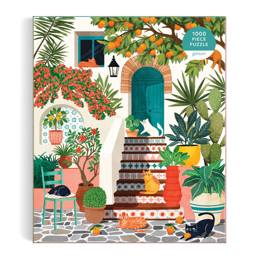 The Tània Garcia "Mediterranean Cats 1000 Piece Puzzle" features a vibrant courtyard scene with lush plants, potted flowers, orange trees, and five cats relaxing on steps and chairs, highlighted by a green door and decorative tiled stairs.