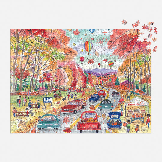 The Michael Storrings Leaf Peeping 1000 Piece Puzzle by Michael Storrings features cars on a tree-lined autumn road, people picnicking and buying food, and hot air balloons drifting overhead—a vibrant fall scene to assemble.