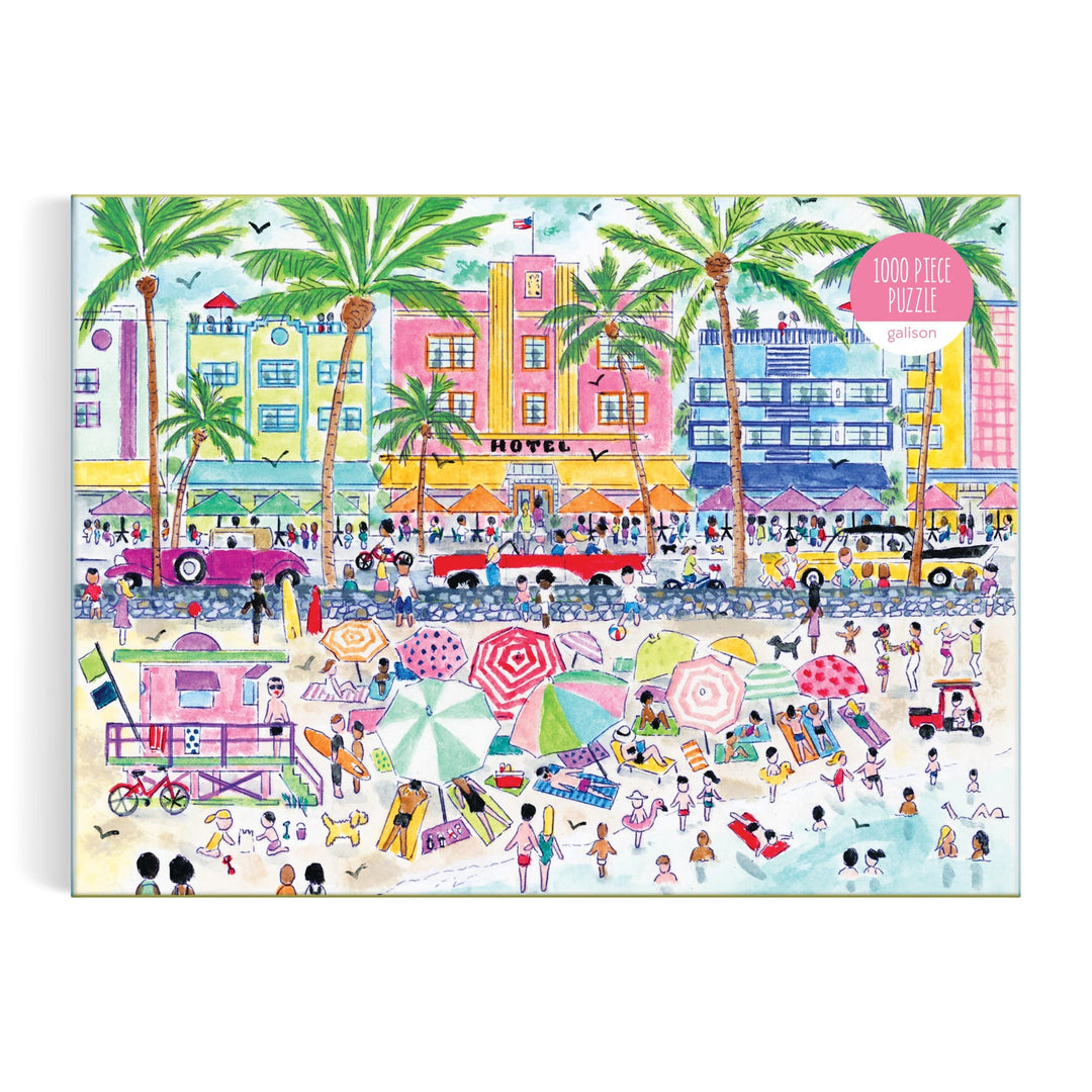 The Michael Storrings South Beach Miami 1000 Piece Puzzle by Michael Storrings features a vibrant illustrated box with sunbathers, umbrellas, palm trees, vintage cars, and Art Deco buildings in a lively beach scene.