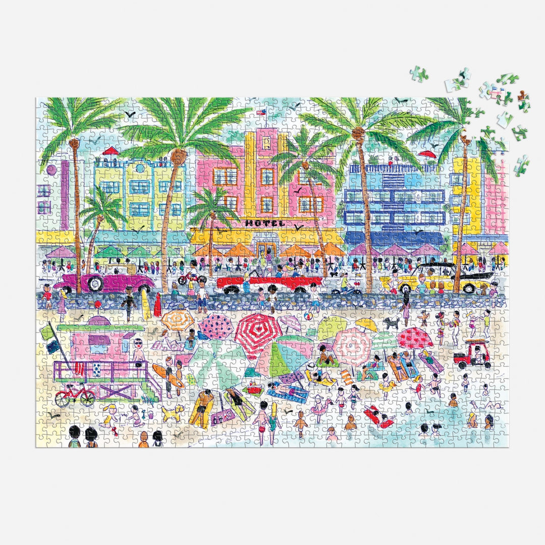 The Michael Storrings South Beach Miami 1000 Piece Puzzle by Michael Storrings features a vibrant beach scene with people, umbrellas, palm trees, and art deco buildings. A few pieces are missing in the top right corner.