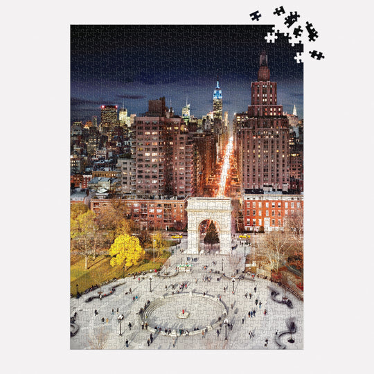 The Stephen Wilkes Day To Night Washington Square Park 1000 Piece Puzzle by Stephen Wilkes shows a vibrant NYC nighttime cityscape with the illuminated arch and skyline; a few pieces are missing in the top right corner.