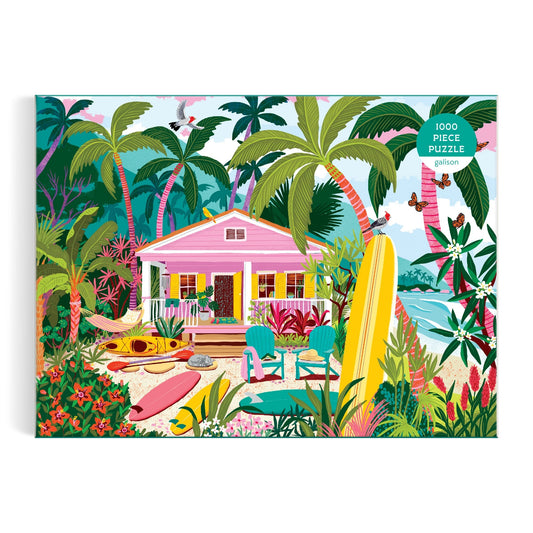 The Surfside Bungalow 1000 Piece Puzzle by Julia Cornelius features a vibrant tropical beach scene with a pink house, surfboards, kayak, palm trees, lush plants, birds, and the ocean on the puzzle box.