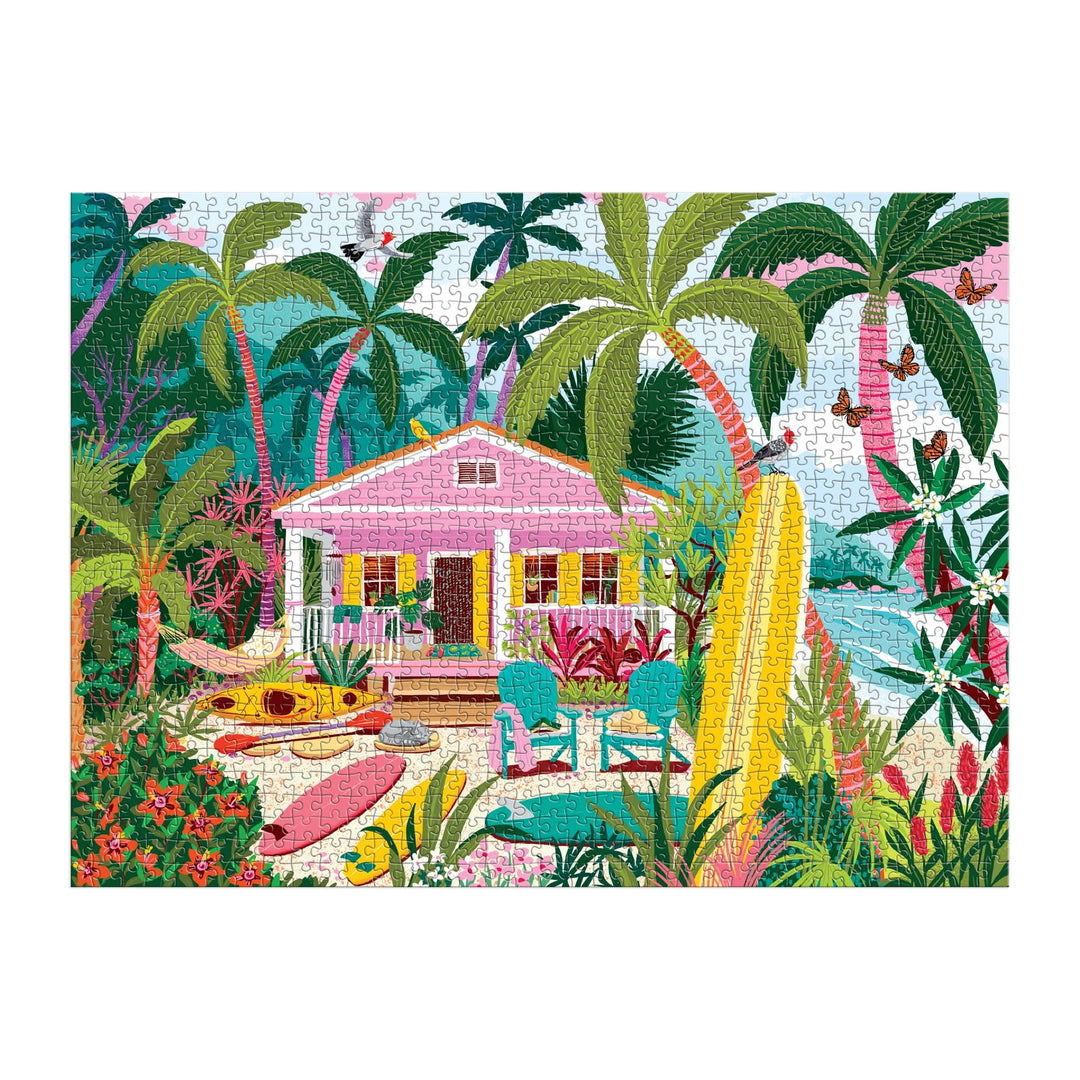 The Surfside Bungalow 1000 Piece Puzzle by Julia Cornelius features a vibrant tropical beach scene with a pink house, palm trees, surfboards, kayak, outdoor chairs, and lush plants by the ocean under a blue sky.