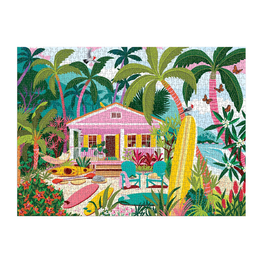 The Surfside Bungalow 1000 Piece Puzzle by Julia Cornelius features a vibrant tropical beach scene with a pink house, palm trees, surfboards, kayak, outdoor chairs, and lush plants by the ocean under a blue sky.