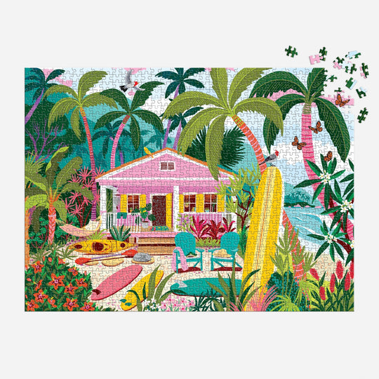 The Surfside Bungalow 1000 Piece Puzzle by Julia Cornelius features a vibrant tropical beach with a pink house, palm trees, surfboards, chairs, and lush plants. The top right corner shows some unfinished pieces for an added challenge.