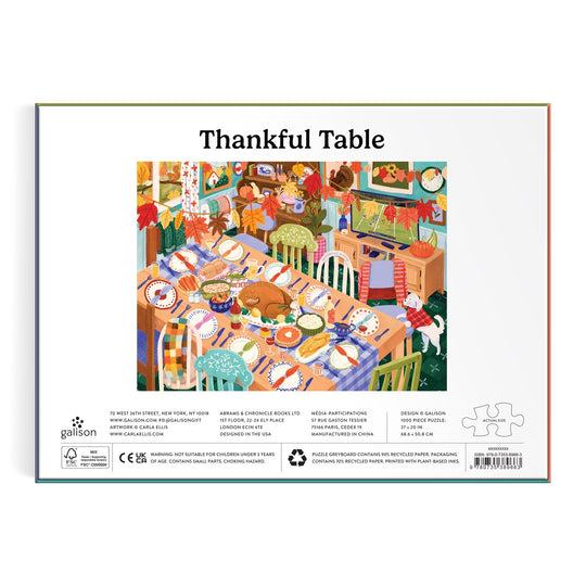 The Carla Ellis Thankful Table 1000 Piece Puzzle features a vibrant illustration of a festive dining room with a long table, mismatched chairs, pumpkins, dishes, and autumn decor. The box is labeled "Thankful Table.