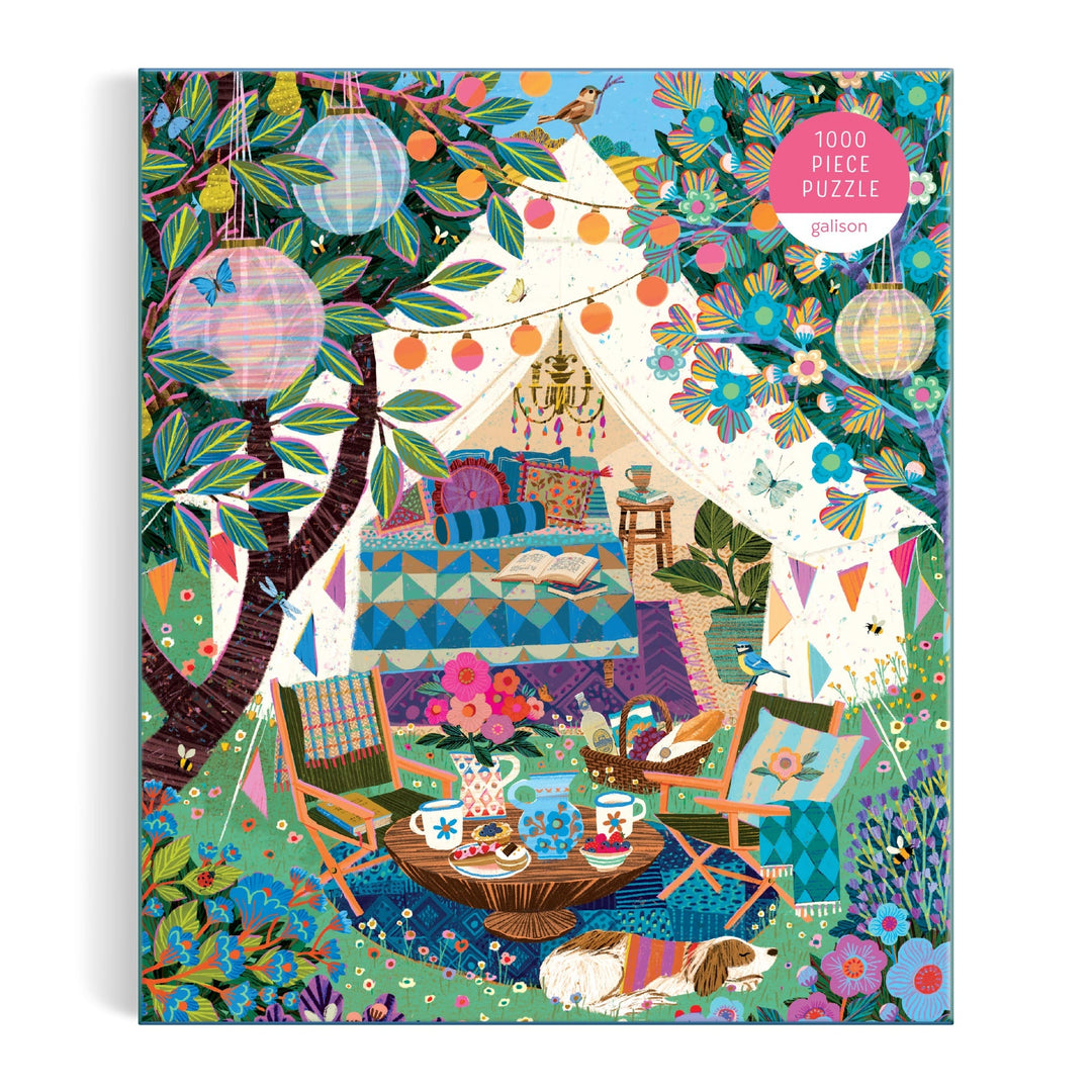The Victoria Ball Blooming Escape 1000 Piece Puzzle by Victoria Ball features a cozy outdoor tent with lanterns and flowers, comfy cushions and quilts inside, plus a tea set, chairs, books, and two small dogs relaxing on the grass.