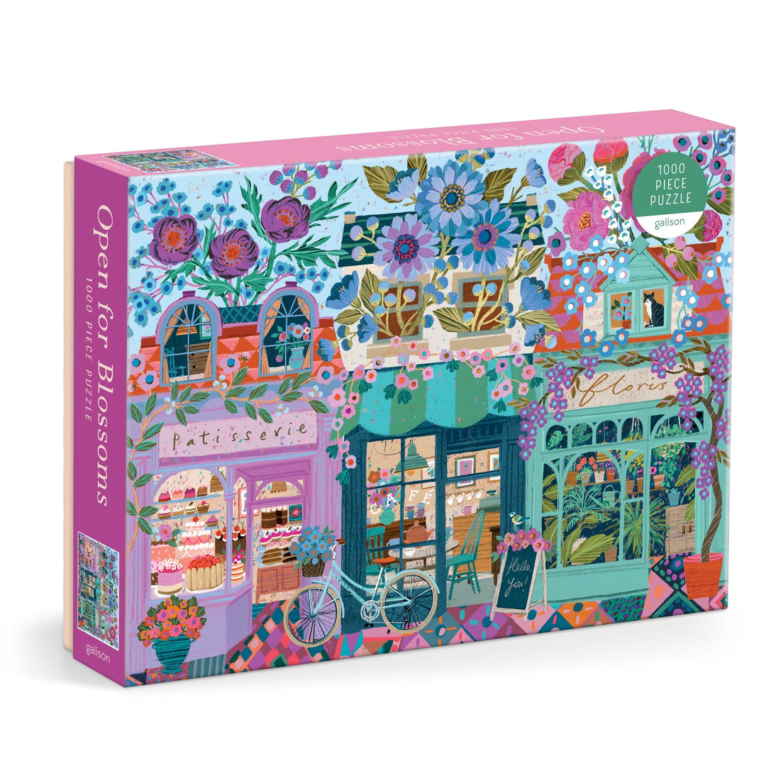 The Victoria Ball Open For Blossoms 1000 Piece Puzzle features a vibrant floral street scene with shops, a bakery, a white bike, and lush blooms. The puzzle box displays the title "Open For Blossoms" and is by Victoria Ball.