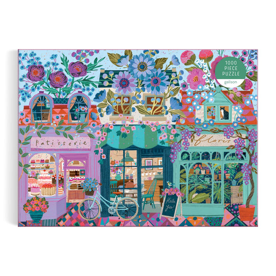 The Victoria Ball Open For Blossoms 1000 Piece Puzzle features a charming street scene with a patisserie, flower shop, café, vibrant flowers and plants, a parked bicycle, and the puzzle’s 1000 piece label in the top right corner.