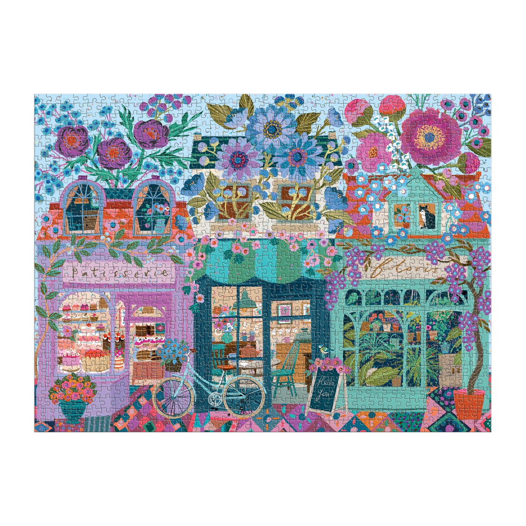 The Victoria Ball Open For Blossoms 1000 Piece Puzzle features a colorful street scene with flower-filled shops, a bakery, a cafe, and a greenhouse. A bicycle rests by the bakery window as large, vibrant flowers brighten the charming setting.