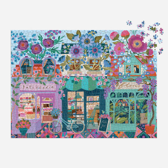 The Victoria Ball Open For Blossoms 1000 Piece Puzzle by Victoria Ball features a vibrant street scene with floral buildings, a bakery window, bicycle, and café. A few pieces are missing from the upper right corner.