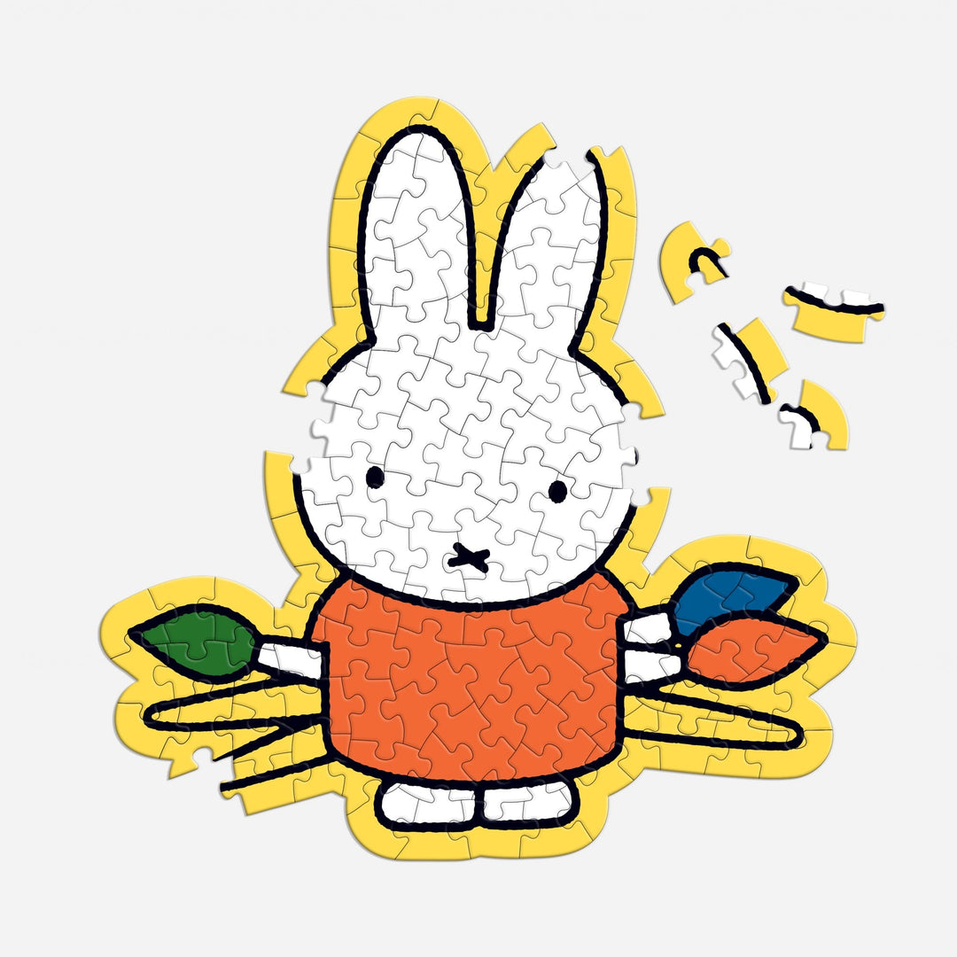 The Miffy 100 Piece Surprise Puzzle by Miffy features a partially completed jigsaw of a white cartoon bunny in an orange shirt, holding green and blue leaves, with missing pieces in the upper right and a yellow outline.
