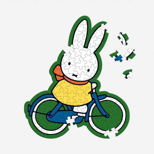 The Miffy 100 Piece Surprise Puzzle by Miffy features a white cartoon rabbit in a yellow shirt and red scarf riding a blue bicycle, with several pieces missing from the upper right corner.