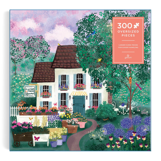 The Joy Laforme Garden Path 300 Piece Puzzle by Joy Laforme features a charming house with a brown roof, vibrant flowers, a garden path, and trees under a whimsical pink and purple sky. A sign reads “Clover & Fern Garden.”.
