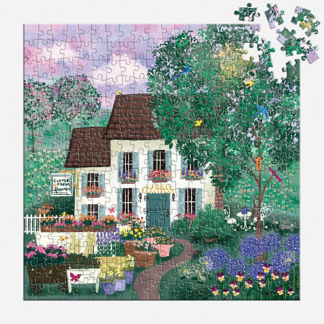 The Joy Laforme Garden Path 300 Piece Puzzle by Joy Laforme features a nearly finished scene of a white cottage with a brown roof amid vibrant flowers and green trees, under a pink and purple sky. A few pieces are missing from the top right corner.