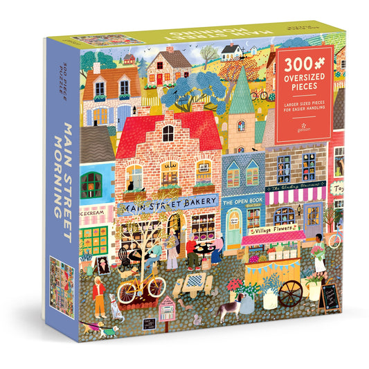 The Elisabeth Dorrian Main Street Morning 300 Piece Puzzle features oversized pieces and a vibrant street scene with shops, a bakery, café, flowers, and people enjoying the day.
