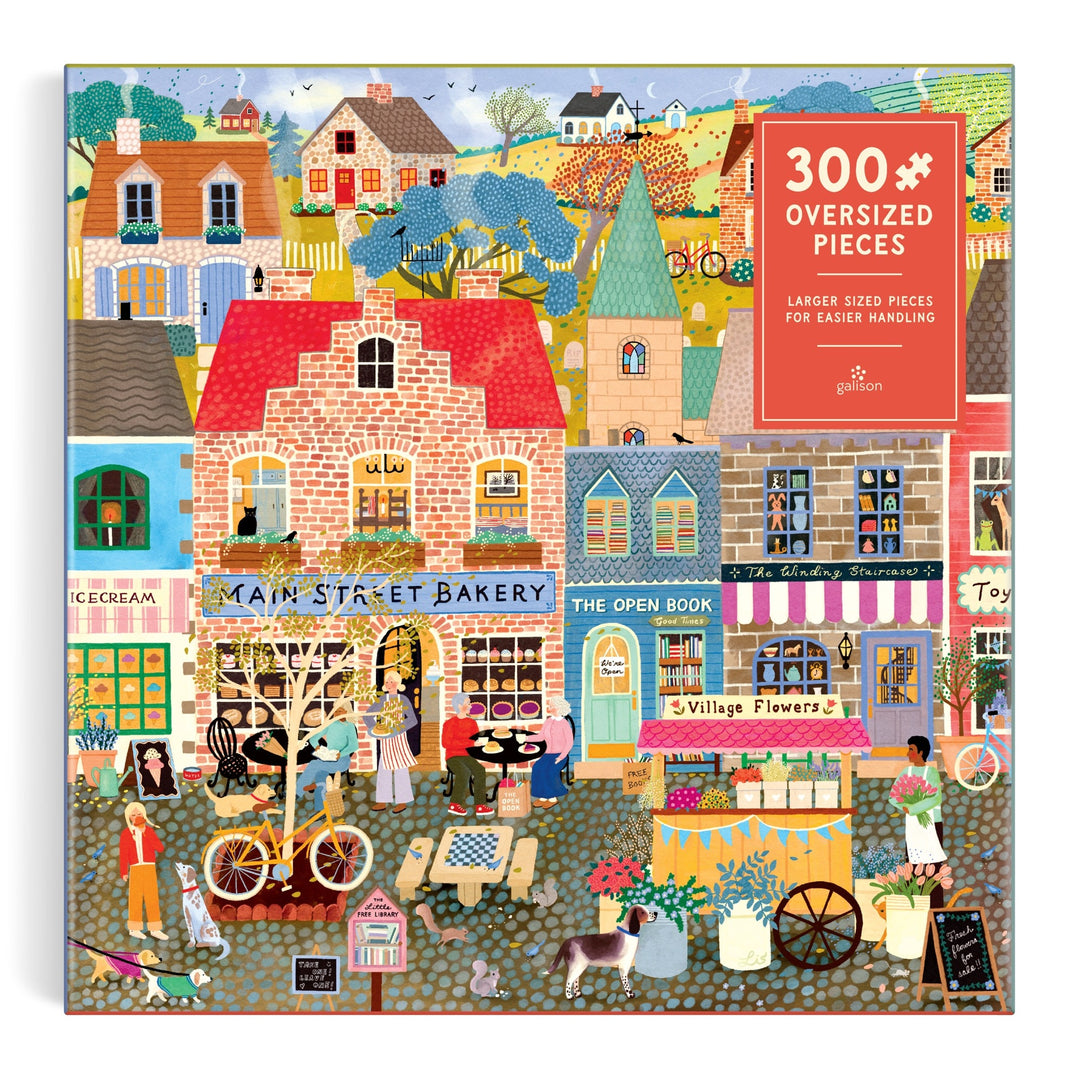 Colorful illustration of a lively village street with shops like a bakery, bookstore, and flower shop, plus people walking and biking; Main Street Morning 300 Piece Puzzle by Elisabeth Dorrian. Puzzle box reads 300 Oversized Pieces.