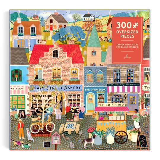 Colorful illustration of a lively village street with shops like a bakery, bookstore, and flower shop, plus people walking and biking; Main Street Morning 300 Piece Puzzle by Elisabeth Dorrian. Puzzle box reads 300 Oversized Pieces.
