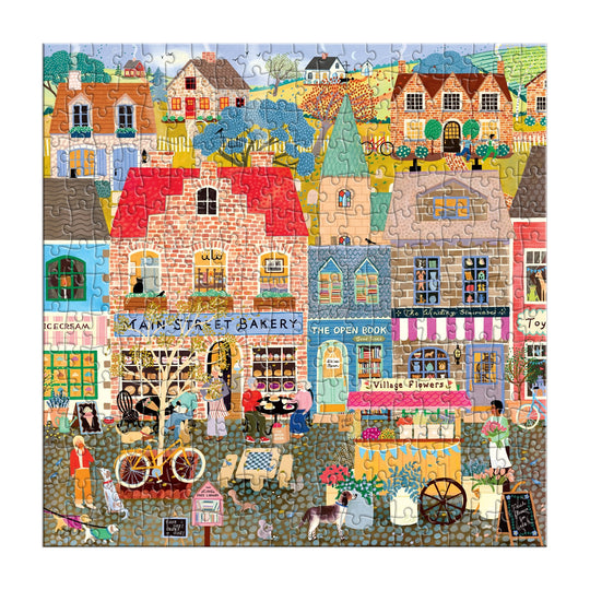 The Main Street Morning 300 Piece Puzzle by Elisabeth Dorrian features a vibrant village scene with shops like a bakery, bookstore, florist, and ice cream parlor, bustling with people, bikes, trees, flowers, and homes nestled among rolling hills.