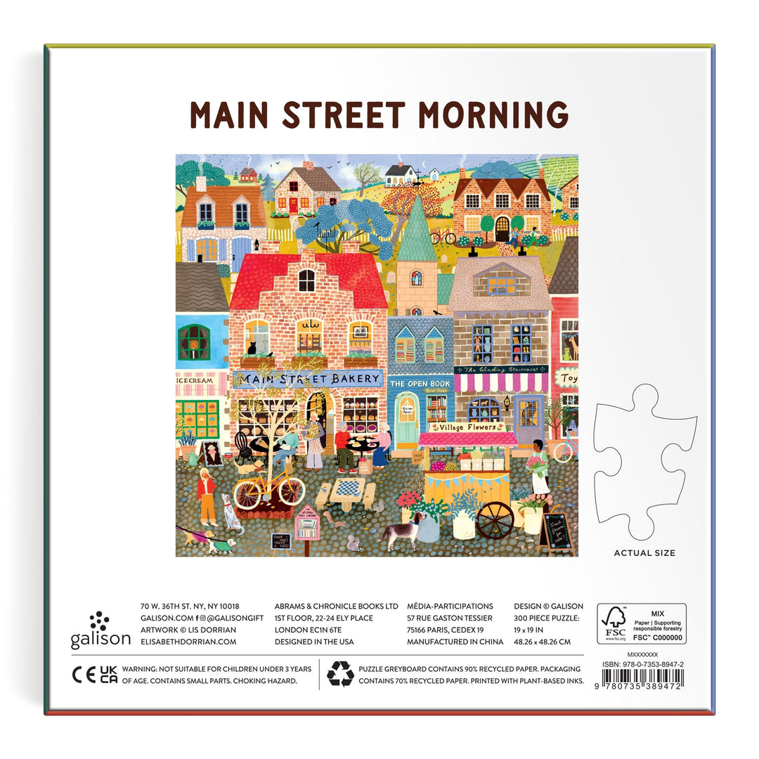 A vibrant illustration of shops, people, bicycles, and dogs in front of quaint buildings appears on the Main Street Morning 300 Piece Puzzle from Elisabeth Dorrian, with a sample puzzle piece and product details displayed on the box.