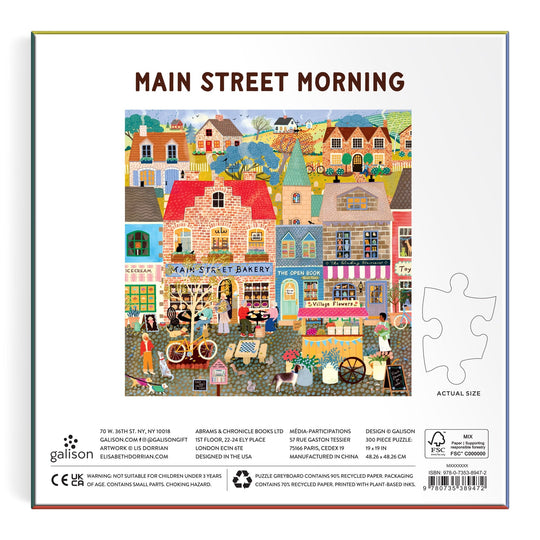 A vibrant illustration of shops, people, bicycles, and dogs in front of quaint buildings appears on the Main Street Morning 300 Piece Puzzle from Elisabeth Dorrian, with a sample puzzle piece and product details displayed on the box.