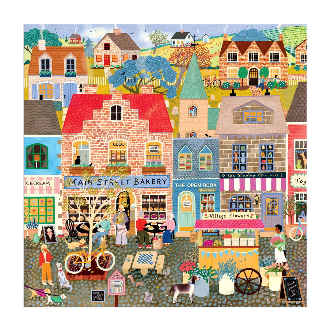 Main Street Morning 300 Piece Puzzle by Elisabeth Dorrian features a vibrant village with shops—a bakery, bookstore, ice cream shop, and flower stand—plus people walking, cycling, chatting, kids playing, a dog, and bright houses with hills in the background.