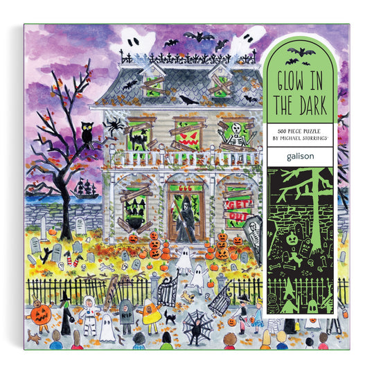 The Michael Storrings Haunted House 500 Piece Glow in the Dark Puzzle features a Halloween scene with ghosts, pumpkins, bats, skeletons, and costumed trick-or-treaters under a purple sky—perfect for intermediate puzzlers.