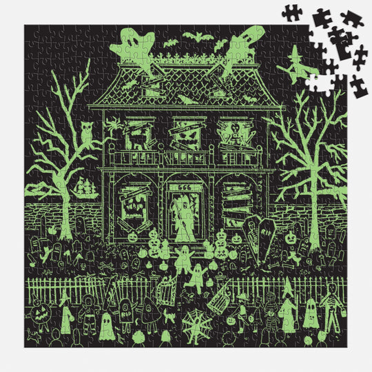 The Michael Storrings Haunted House 500 Piece Glow in the Dark Puzzle, by Michael Storrings, depicts a haunted house with ghosts, skeletons, eerie trees, and gravestones. A few pieces are missing from the upper right; the image glows green in the dark.